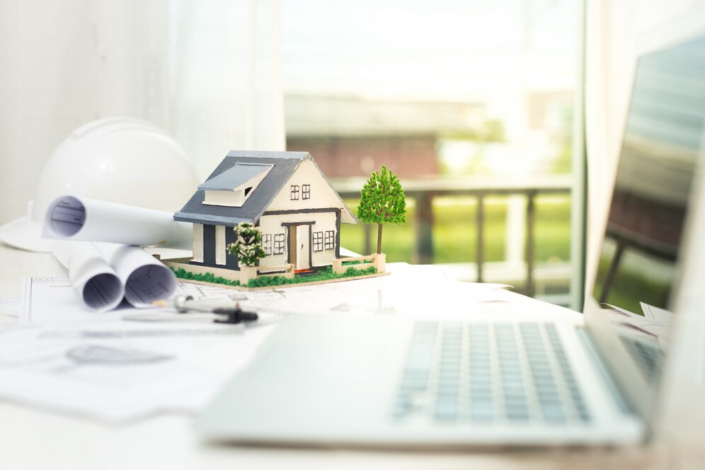 op Real Estate Agent in Mission Viejo working on property plans with a model house, blueprints, and a laptop on the desk